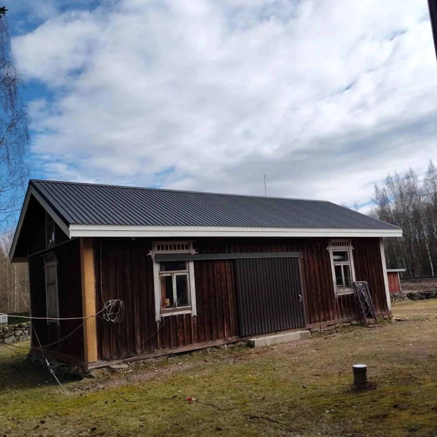 Vanha, tummanruskea puurakennus, jossa on tummanharmaa aaltopeltikatto ja valkoiset räystäslaudat. Rakennuksen seinissä on useita valkoisia ikkunoita ja suuri tummanruskea liukuovi. Maassa on ruohoa ja sammalta. Taustalla näkyy puita ja sininen taivas valkoisilla pilvillä. Kuva on otettu päivänvalossa.