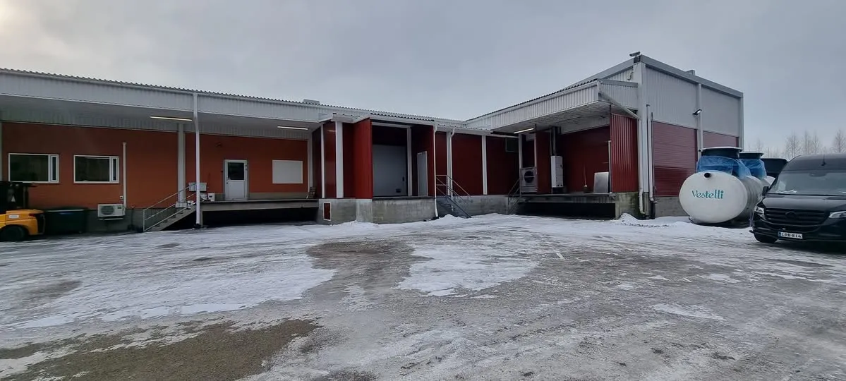 Kuva teollisuusrakennuksesta tai varastosta talvisena, pilvisenä päivänä. Rakennuksen seinät ovat oranssinpunaiset ja tummanpunaiset, ja siinä on useita lastauslaitureita. Maassa on lunta ja jäätä. Vasemmalla näkyy keltainen trukki ja oikealla musta pakettiauto. Rakennuksen edessä on kaksi valkoista säiliötä, joista toisessa on Vestelli-logo.